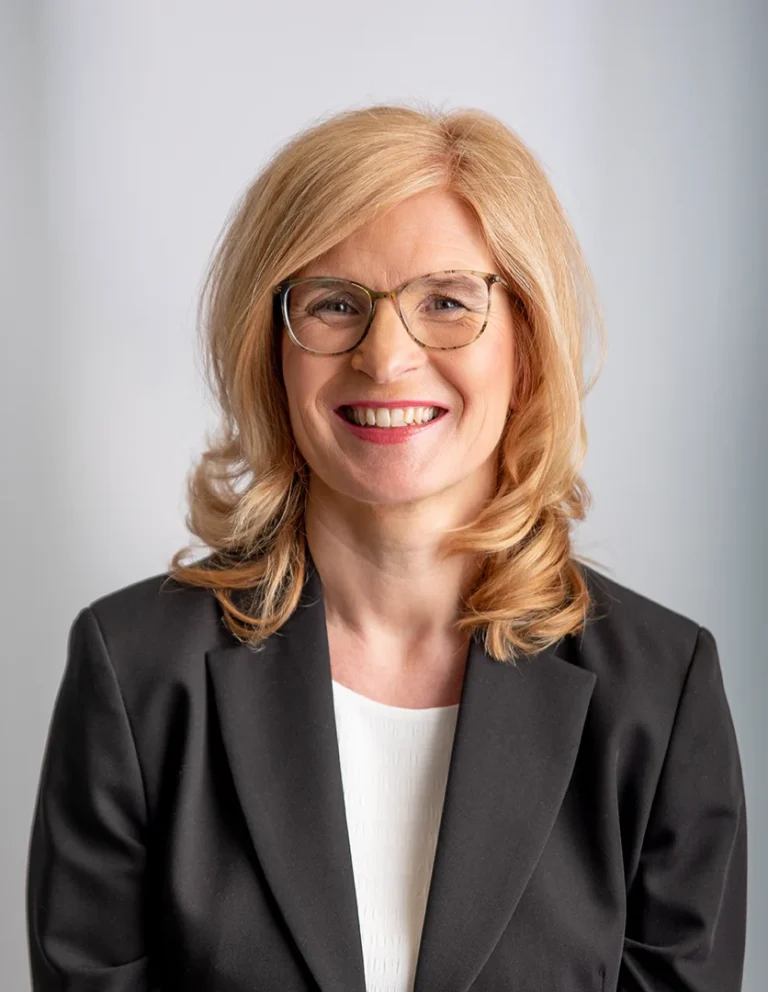 Frau mit blonden, gewellten Haaren und Brille, trägt ein gestreift blaues Hemd.